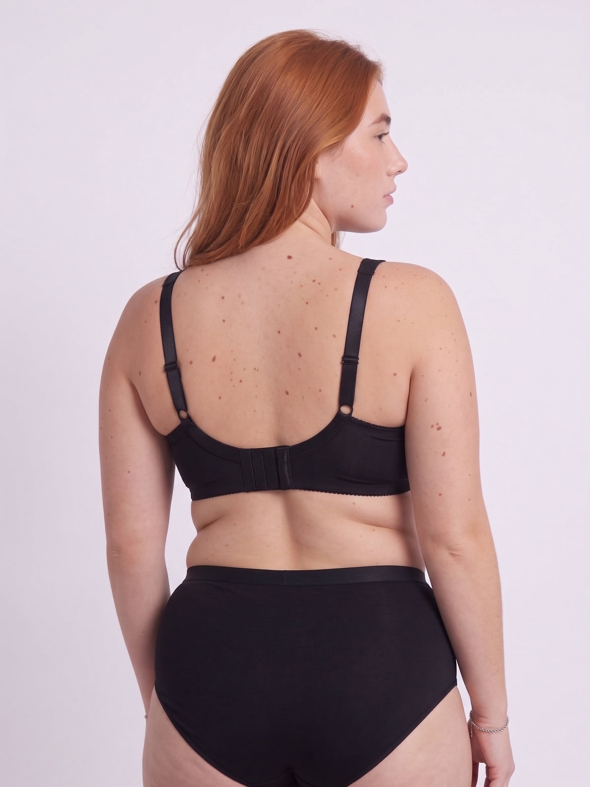 Back view of a curvy red-haired model wearing a black wireless bra and matching panties. The image highlights the wide, adjustable back band, demonstrating how a properly fitted band provides 80% of the support for a large bust, effectively relieving shoulder strain without the use of rigid underwires.