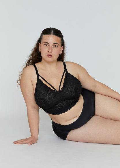 A seated plus-size model wearing the black Mia wireless lace bralette for large breasts, featuring strappy cleavage details and high-waisted briefs in a comfortable pose