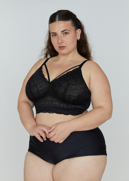 Full-body portrait of the plus-size model with sleek dark hair from image_9.png, standing in a studio wearing the black wire-free Mia lace bralette with its decorative cross-strap cleavage and wide lace band. She wears matching high-waisted black briefs, hands clasped, showing the full fit and lace details against an off-white background. Her tattoos are visible.