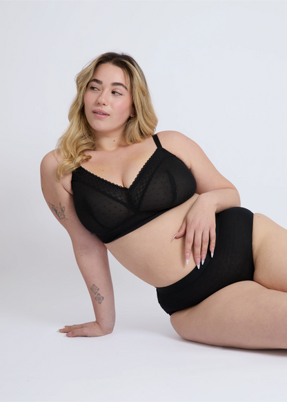 Plus-size model with tattoos, lying down in a supportive pose, wearing the NewG Alma black wireless bra for large busts, showcasing the dotted mesh and lace details.