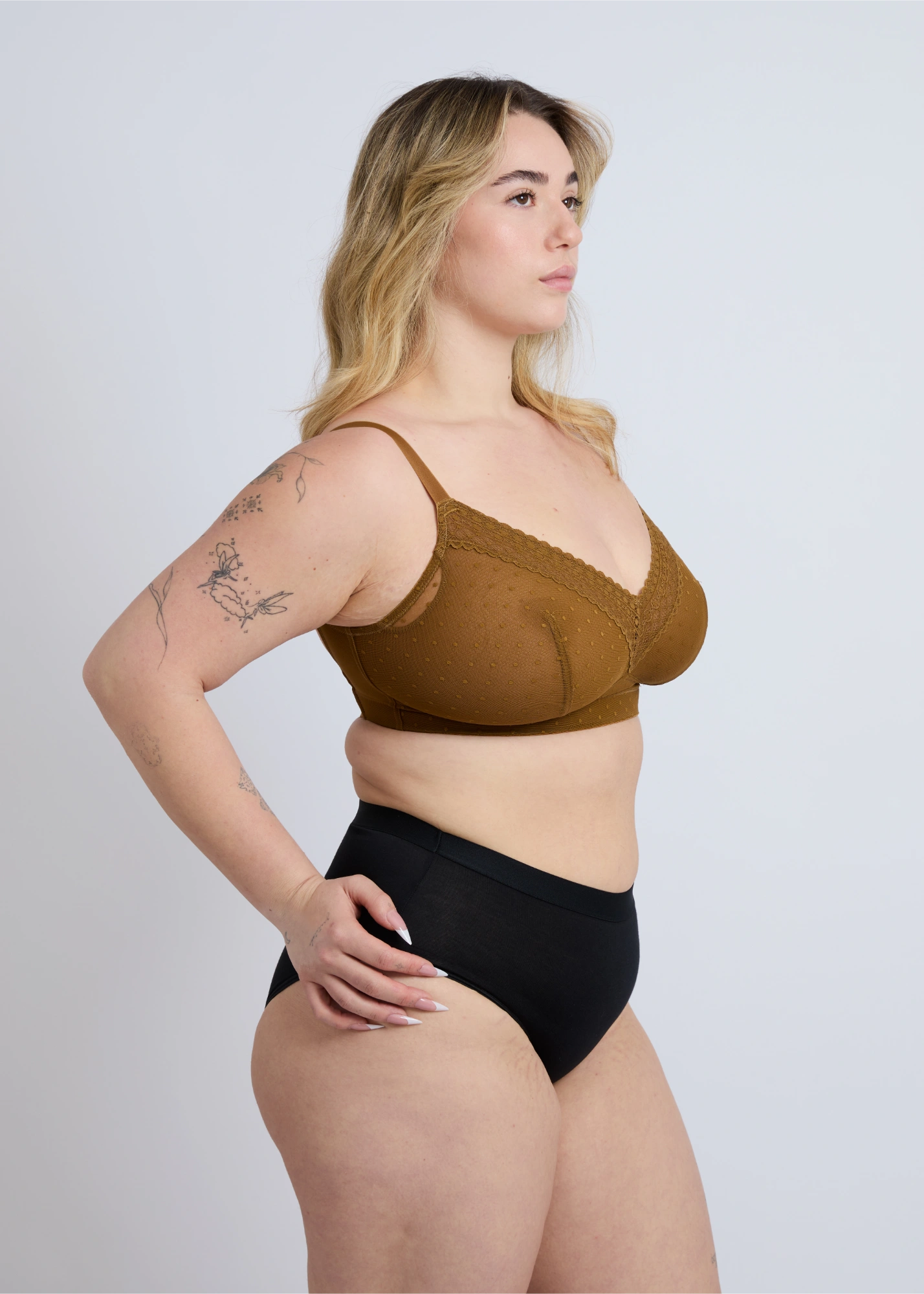 A three-quarter side profile view of a plus-size blonde model wearing the NewG Alma bronze-gold lace wire-free bra for large busts, paired with black briefs, against a white studio background, showcasing the fit and dotted mesh pattern.