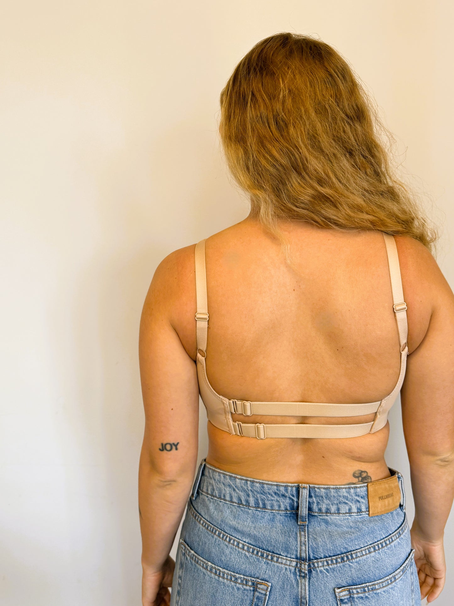 Rear lifestyle view of the NewG Doni beige wireless bra for large busts, showing the low back double strap design on a model with long hair and denim jeans.