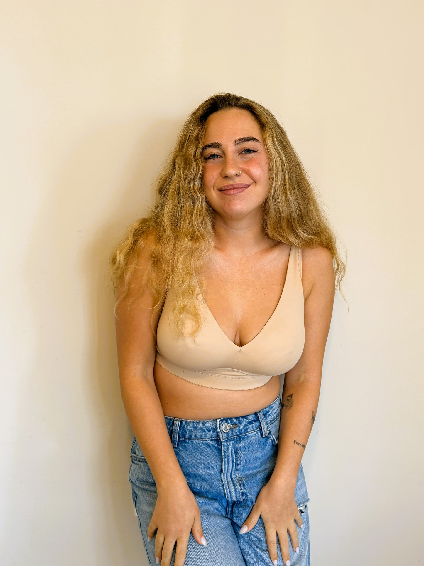 A smiling lifestyle photo of a model with a large bust wearing the NewG Doni beige wireless bra and jeans, highlighting comfortable daily support and a natural fit.